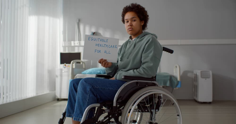 Distressed African-American woman in a hospital wheelchair holds a "Equitable Health Care for All" poster.