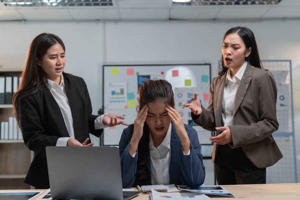 worker at desk in distress surrounded by angry coworkers