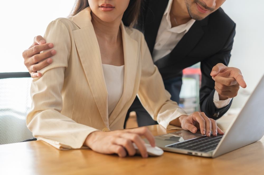 Uncomfortable Asian woman at work looking at a male colleague’s hand on her shoulder, showing signs of distress and discomfort due to unwanted physical contact.