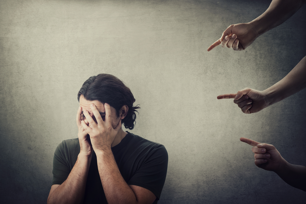 Man covering his face with his hands, looking distressed, as multiple hands point at him from behind, symbolizing blame, shame, and emotional pressure.