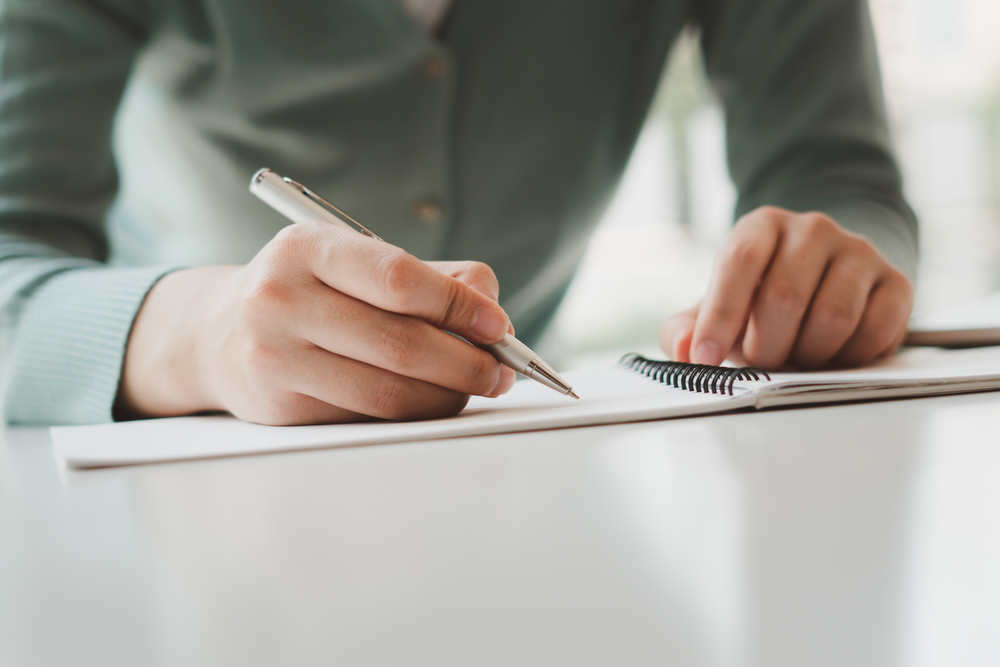 Businesswoman writing on a notepad with a pen while reviewing financial statements in an office.