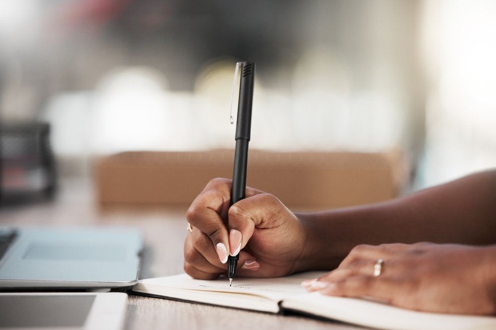 Close-up of hands writing in a notebook at an office desk, planning a project schedule with notes on deadlines, feedback, and development ideas.