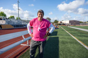 Person standing at a track field wearing a pink breast cancer awareness T-shirt smiling into the camera