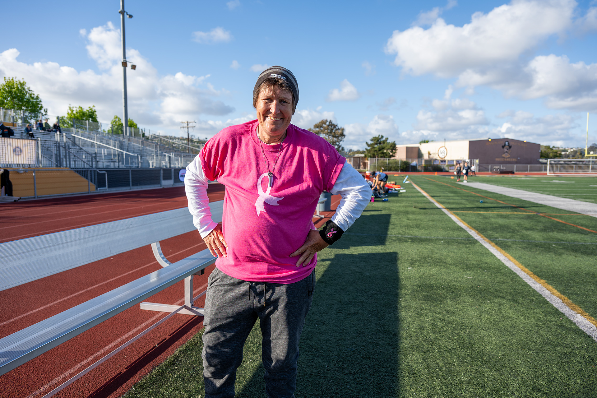 Person standing at a track field wearing a pink breast cancer awareness T-shirt smiling into the camera
