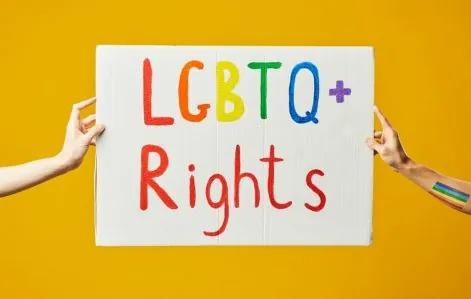 Vibrant photo of two hands holding an LGBTQ+ rights sign against a yellow background.